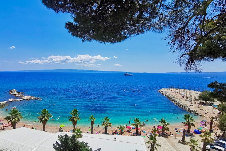 Vista dall'alto di una spiaggia assolata con persone che nuotano in acqua turchese e si rilassano sulla sabbia costeggiata da palme.
