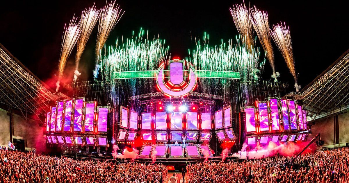 Un'ampia inquadratura di un festival musicale notturno, con una folla immensa di fronte a un palco illuminato da luci rosa e fuochi d'artificio.