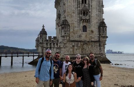 Eine WeRoad-Gruppenreise von sieben Personen posiert an einem Sandstrand vor einem historischen Steinturm am Wasser.