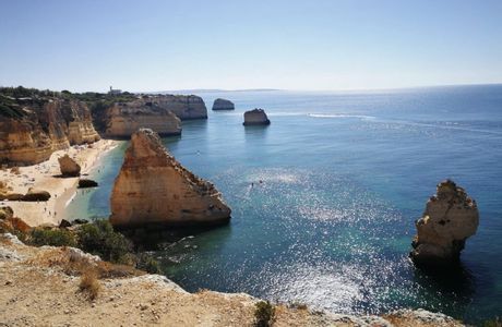 Erhöhter Blick auf eine goldene Felsenküste mit einem Sandstrand und großen Felsformationen in einem funkelnden blauen Meer.