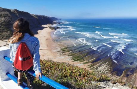 Eine Frau mit einem roten WeRoad Rucksack sitzt auf einem blauen Geländer und blickt auf eine sonnige Küste mit Sandstrand und Klippen.