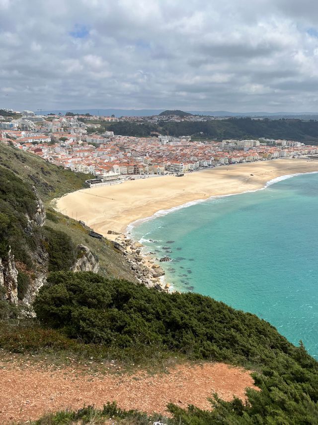 Eine Vogelperspektive auf eine Küstenstadt mit rot gedeckten Dächern, eingebettet zwischen einem breiten Sandstrand und türkisblauem Wasser.
