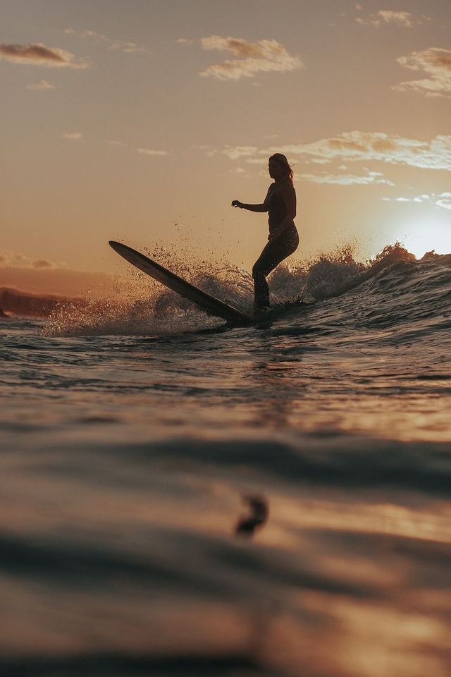 Ein Surfer gleitet als Silhouette auf einer Welle, während die Sonne über dem Meer untergeht.