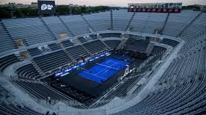 Vista dall'alto di un campo da padel blu illuminato, situato al centro di un grande anfiteatro vuoto in pietra al tramonto.