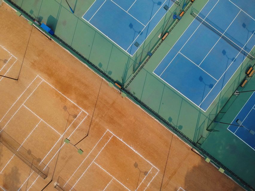 Veduta aerea di campi da tennis vuoti, con campi in terra battuta marroni accanto a campi in cemento blu e verdi.