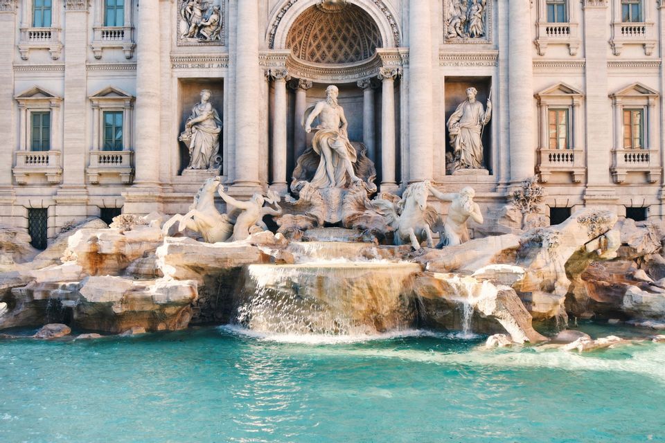 Una grande fontana in marmo ornata con statue classiche, con acqua che scende in una piscina turchese davanti a un imponente edificio.