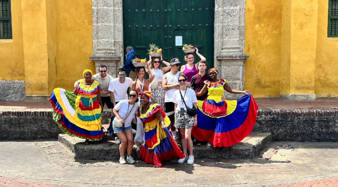 Un gruppo di viaggio WeRoad posa per una foto su gradini di pietra con donne che indossano abiti tradizionali colorati di fronte a un edificio giallo.