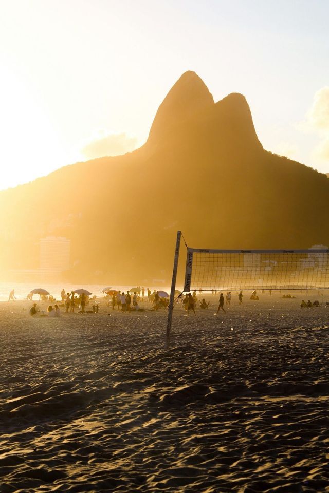 Una rete da pallavolo si erge su una spiaggia affollata al tramonto, con una grande montagna che si staglia contro la luce dorata.