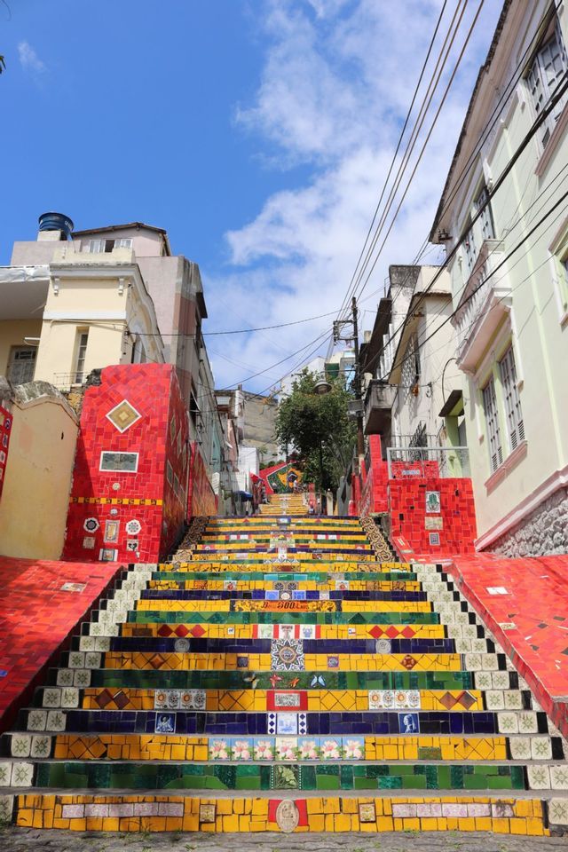 Una ripida scala esterna coperta da colorate piastrelle a mosaico rosse, gialle e blu sale tra gli edifici sotto un cielo blu.