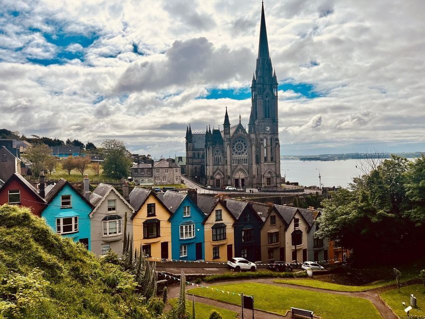 Una hilera de casas coloridas se asienta debajo de una gran catedral gótica en una ladera con vistas a un cuerpo de agua bajo un cielo nublado.