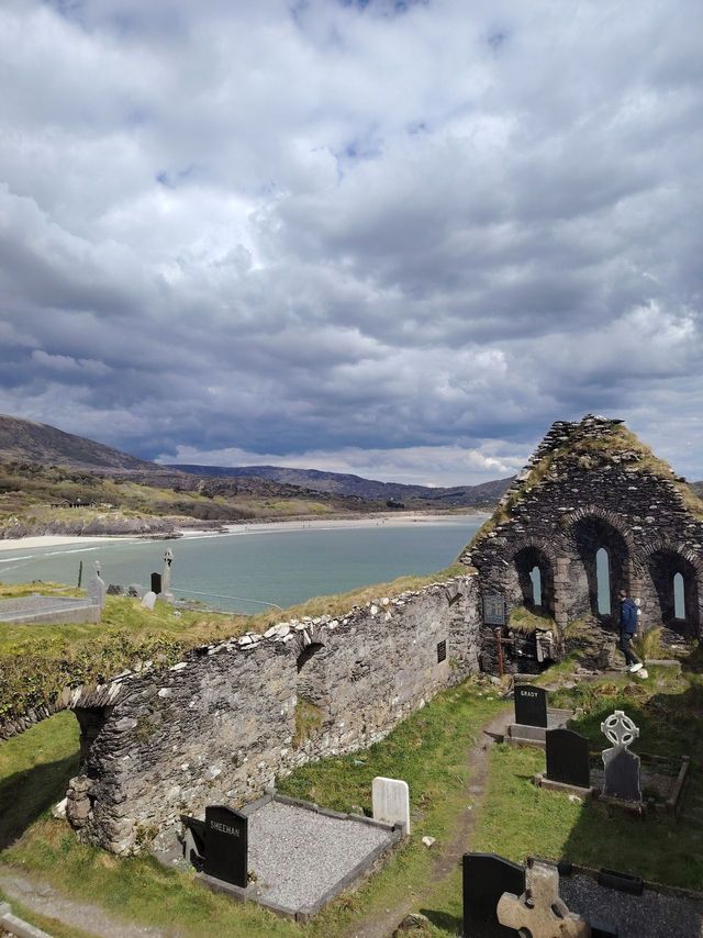 Ruinas de piedra de una iglesia y un cementerio con vistas a una bahía costera y montañas bajo un cielo nublado.