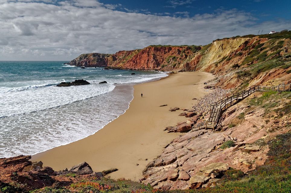 Eine Person geht an einem breiten Sandstrand am Meer entlang, mit rötlichen Klippen und einer Holztreppe im Hintergrund.