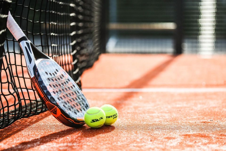 Una racchetta da padel appoggiata alla rete con due palline gialle su un campo arancione.