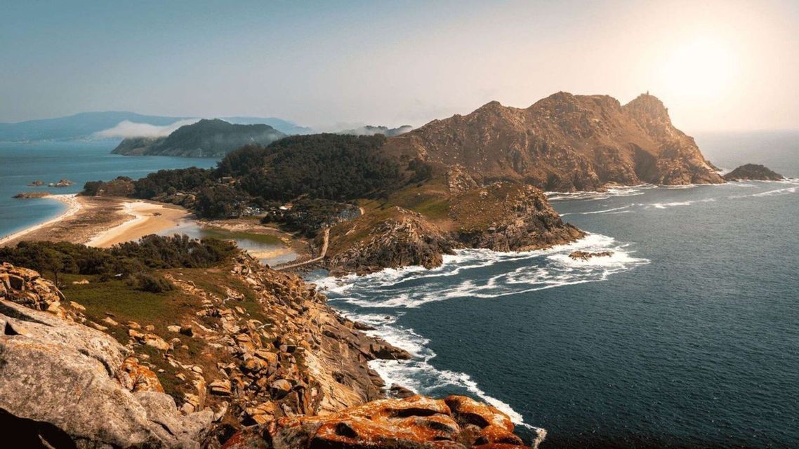 Una vista en picada de una península rocosa y arbolada que incluye un istmo de arena y una pequeña laguna, rodeada por el intenso azul del mar.