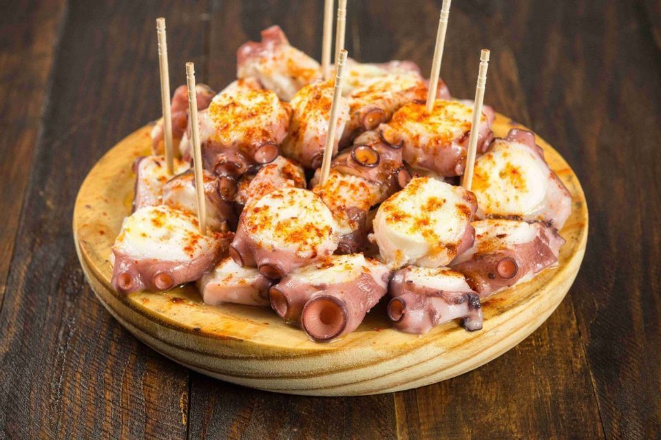 Trozos de pulpo cocido sazonados con pimentón, servidos con palillos en un plato de madera redondo.