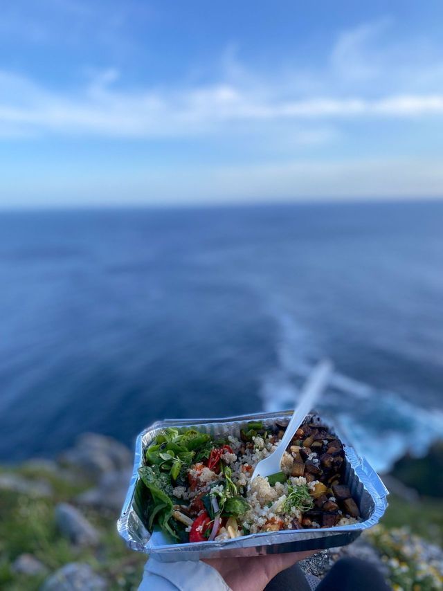 Una persona sosteniendo un recipiente de ensalada para llevar con un tenedor, con el océano azul de fondo.