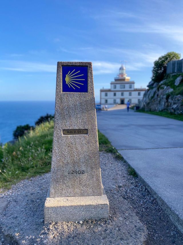 Un hito del Camino de Santiago en Galicia marca el 'Km 0.000' en un sendero costero, con un faro y el océano de fondo.