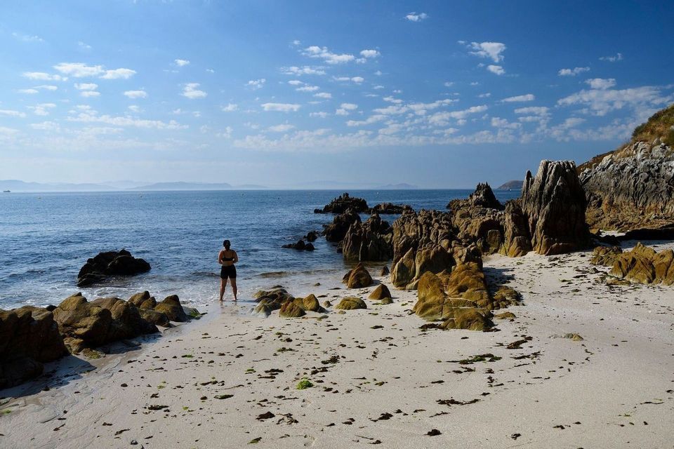 Una persona se encuentra a la orilla del agua en una playa de arena y rocas, contemplando el océano bajo un cielo azul con nubes dispersas.