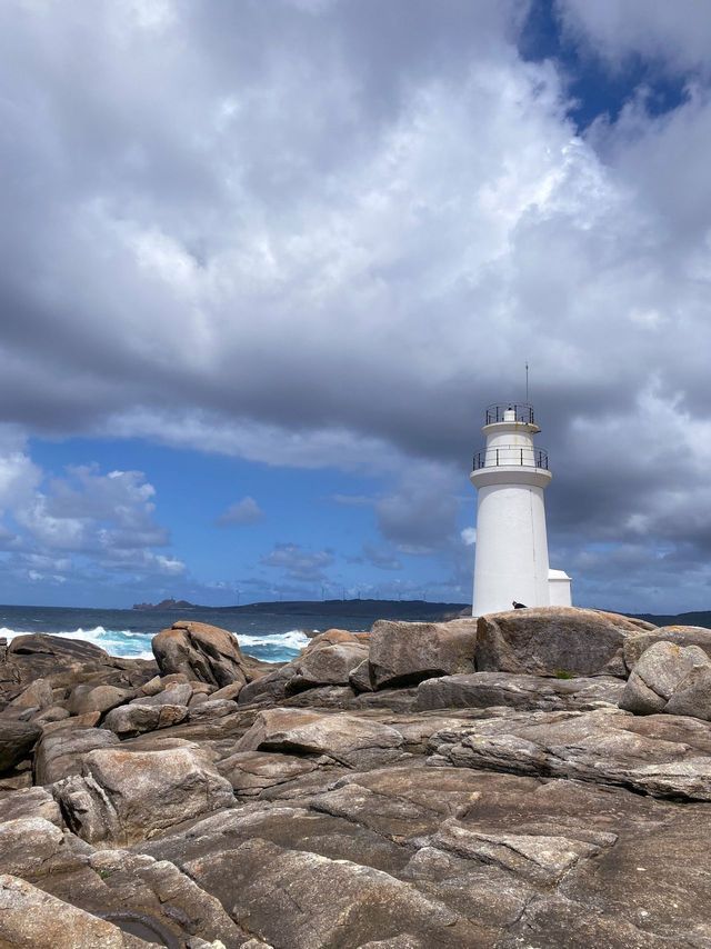 Un faro blanco se alza en una costa rocosa junto a un mar agitado bajo un cielo lleno de dramáticas nubes grises y blancas.