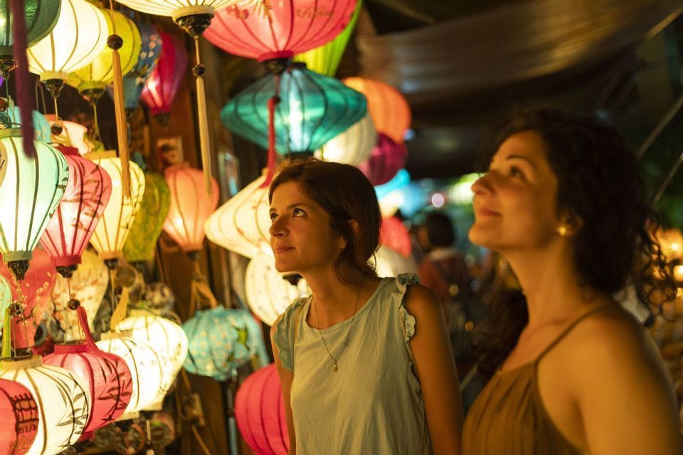 Two women stand at a market stall at night, looking up at a display of colorful, illuminated paper lanterns.