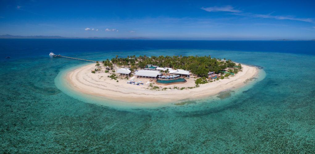 Vista aerea di una piccola isola tropicale con un resort, circondata da una spiaggia di sabbia bianca e acqua turchese cristallina.