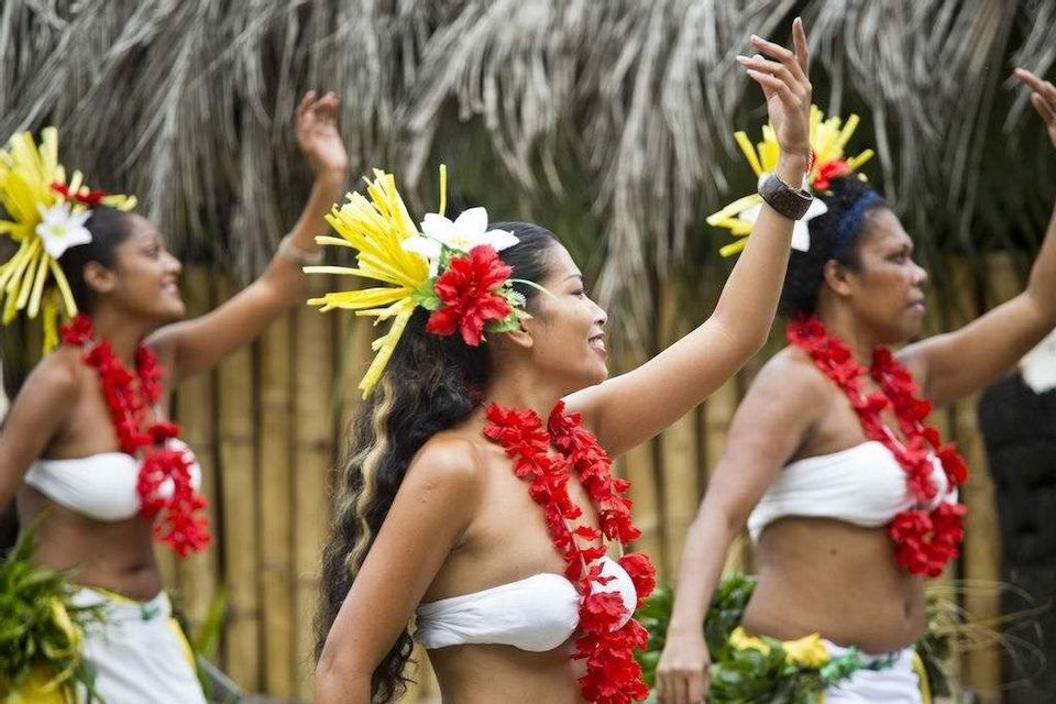 Tre donne di profilo eseguono una danza tradizionale, indossando ghirlande di fiori rossi e copricapi floreali colorati.