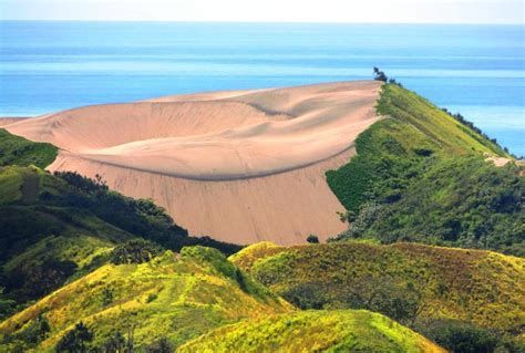Un'ampia duna di sabbia si erge accanto a verdi colline lussureggianti, con l'oceano blu sullo sfondo.