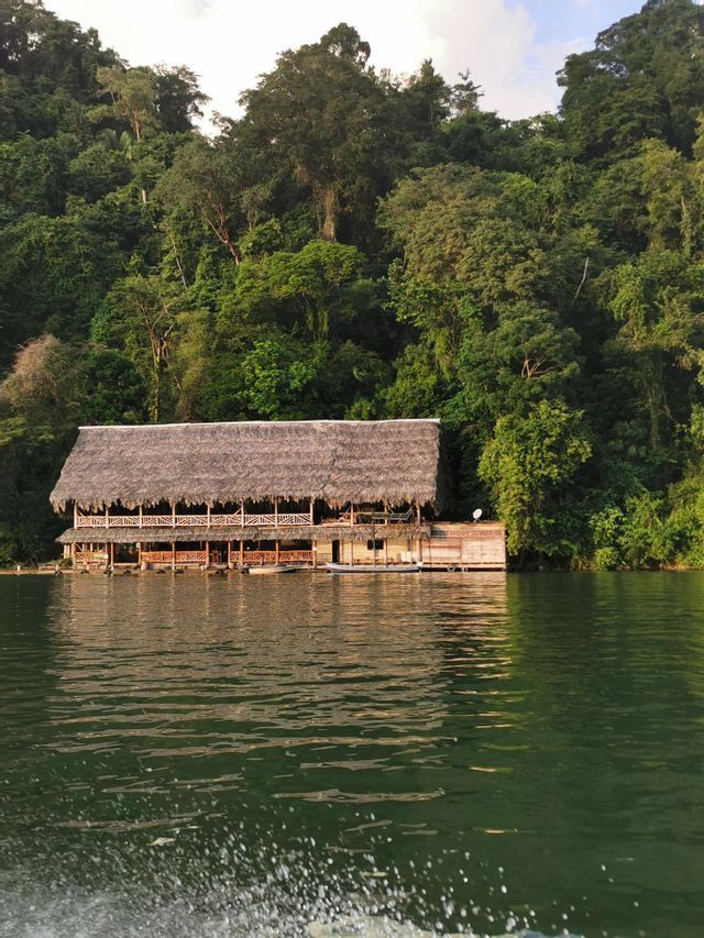 Un lodge en bois à deux étages, avec un toit de chaume, se dresse sur l'eau au pied d'une colline dense et boisée.