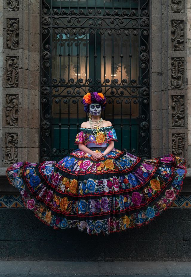 Eine Frau mit Calavera-Gesichtsbemalung und einem Blumenkranz sitzt in einem farbenfrohen, gestuften, bestickten Kleid auf einem Steinsims.