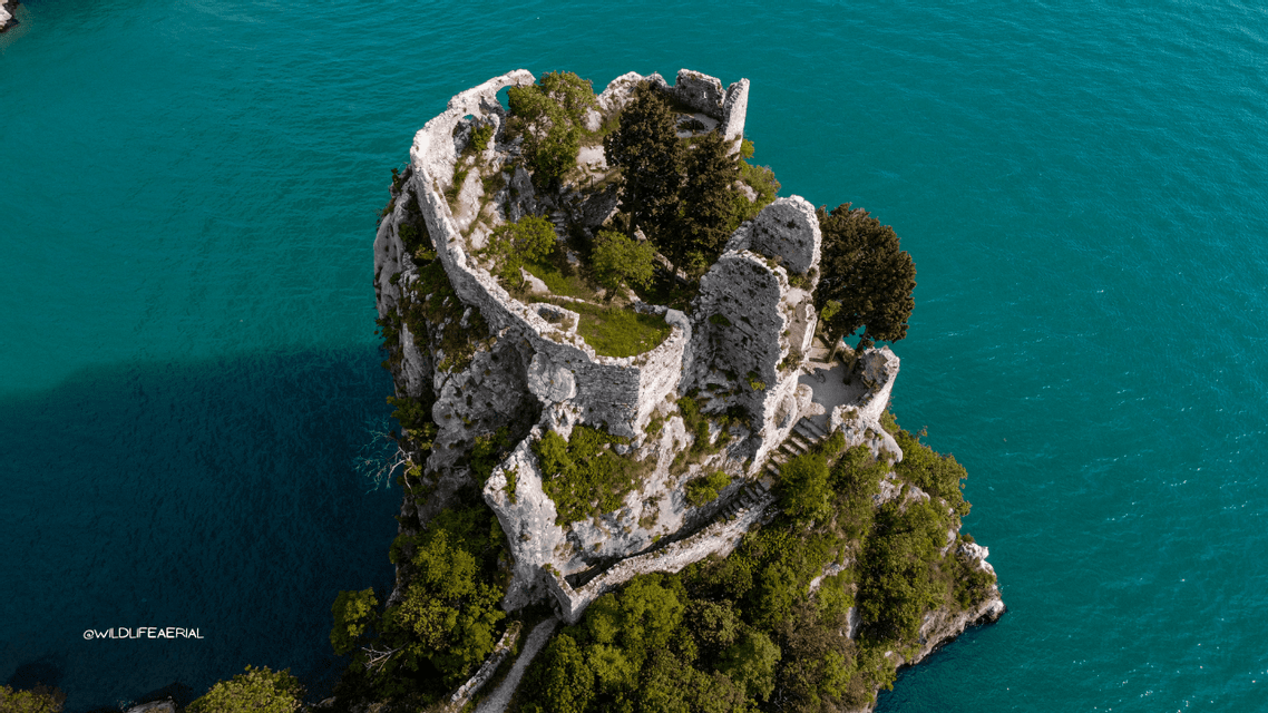 Una vista aerea di rovine di un castello in pietra su un'isola rocciosa coperta da alberi, circondata da acqua turchese.