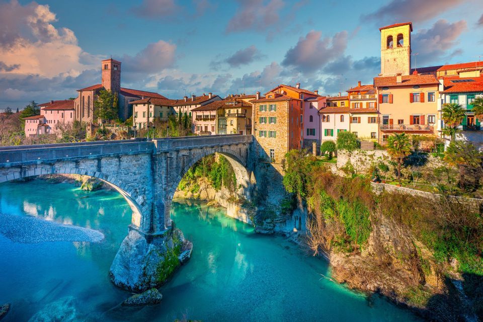 Un ponte in pietra ad arco attraversa un fiume turchese, che conduce a una colorata città collinare con edifici storici sotto un cielo parzialmente nuvoloso.