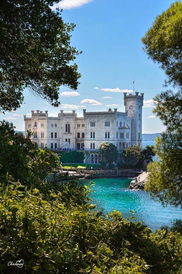 Un castello di pietra bianca si erge su una costa rocciosa accanto ad acque turchesi, visto attraverso alberi verdi lussureggianti sotto un cielo parzialmente nuvoloso.