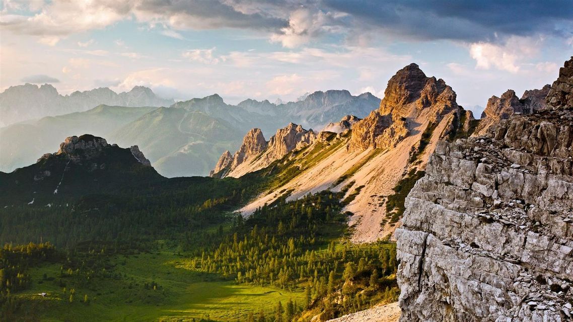 Un'ampia vista di cime montuose rocciose illuminate dal sole che sovrastano valli verdi e fitte foreste sotto un cielo nuvoloso.