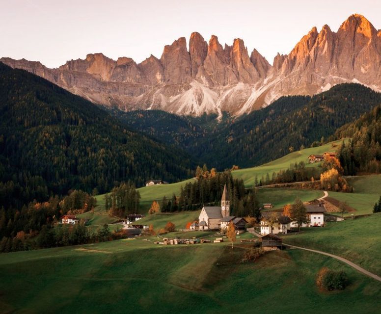 Un piccolo villaggio con una chiesa è incastonato tra dolci colline verdi, alla base di una catena montuosa frastagliata e illuminata dal sole.