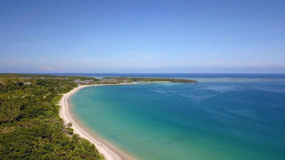 Veduta aerea di una spiaggia curva di sabbia bianca, che costeggia acqua turchese e una lussureggiante foresta verde, sotto un cielo sereno.