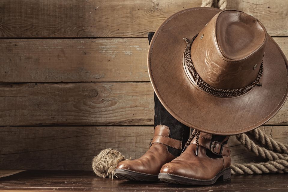 A brown cowboy hat rests on a pair of leather boots, set against a background of wooden planks with a rope.
