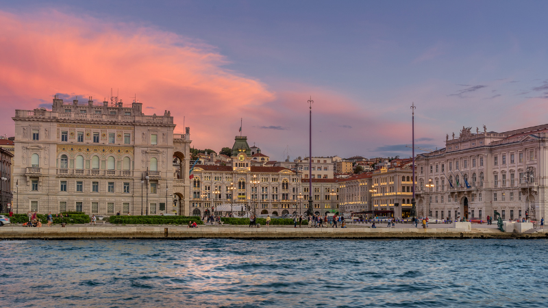 Una vista sul lungomare di sontuosi edifici cittadini illuminati al tramonto sotto un cielo rosa e viola.