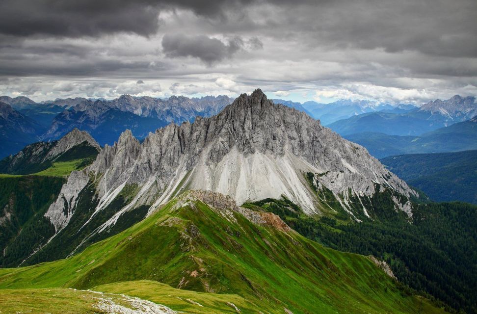 Una vetta montuosa frastagliata e rocciosa si erge dietro un crinale verde e erboso sotto un cielo nuvoloso.