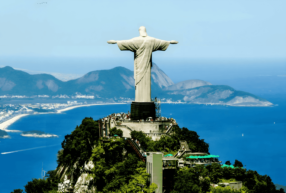 Une grande statue aux bras tendus se dresse sur un sommet de montagne verdoyant, dominant un littoral étendu et une vaste baie bleue.