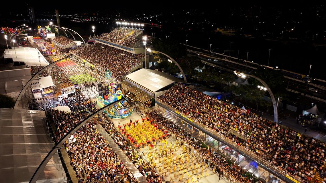 Vista aérea de un vibrante desfile de carnaval nocturno con artistas disfrazados y carrozas elaboradas, rodeado de grandes multitudes en las gradas.