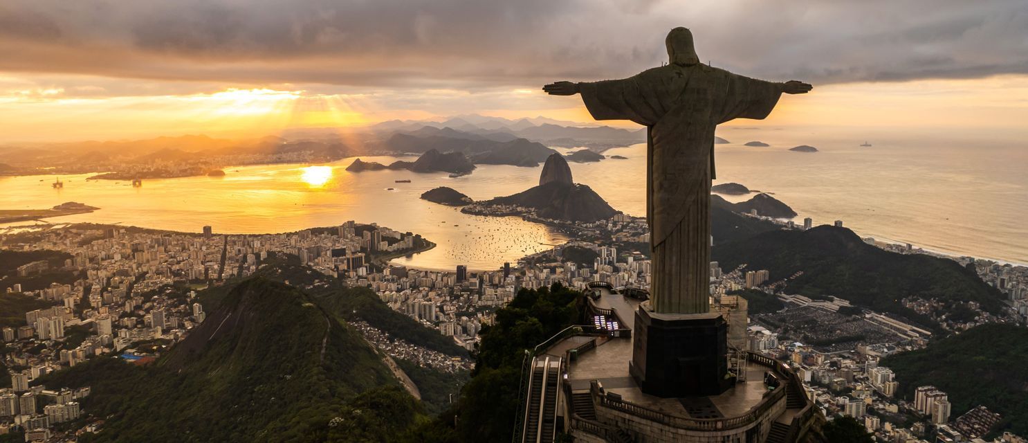 La estatua del Cristo Redentor domina una ciudad costera, una bahía y montañas durante un atardecer dorado.