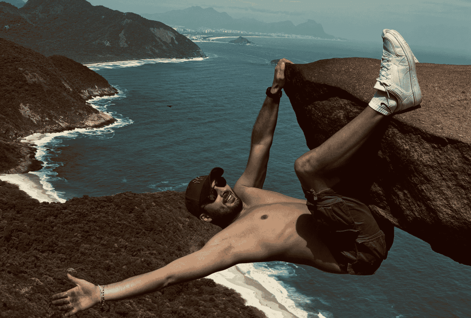 Un homme souriant suspendu à un rocher pour une photo, avec une vue plongeante sur une côte avec des plages et des collines luxuriantes.