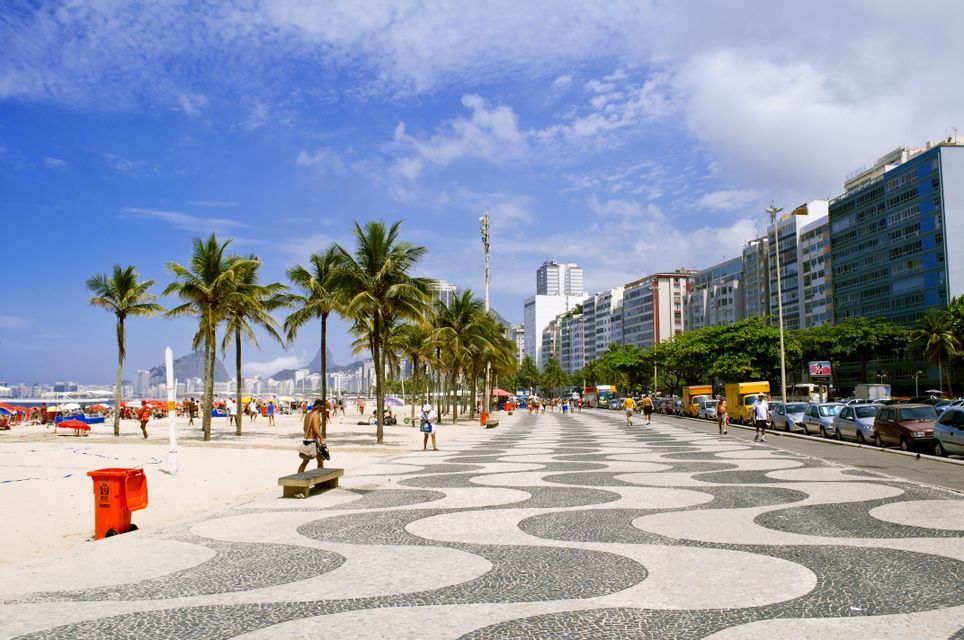 Un lungomare in mosaico con motivi a onda costeggia una spiaggia sabbiosa fiancheggiata da palme, con lo skyline di una città sullo sfondo.