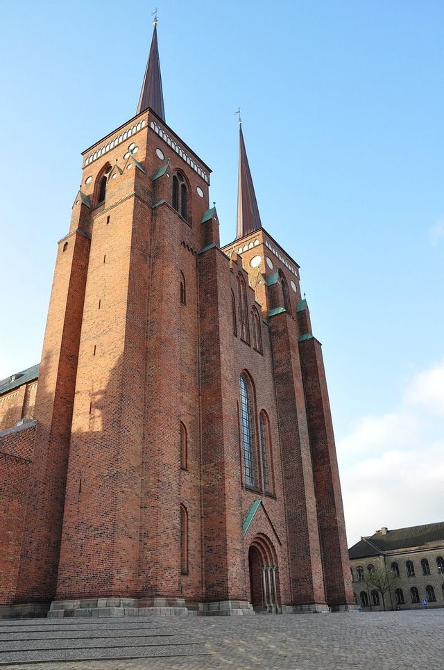 Eine Untersicht auf eine große Kathedrale aus rotem Backstein mit zwei Türmen und hohen Turmspitzen vor blauem Himmel.