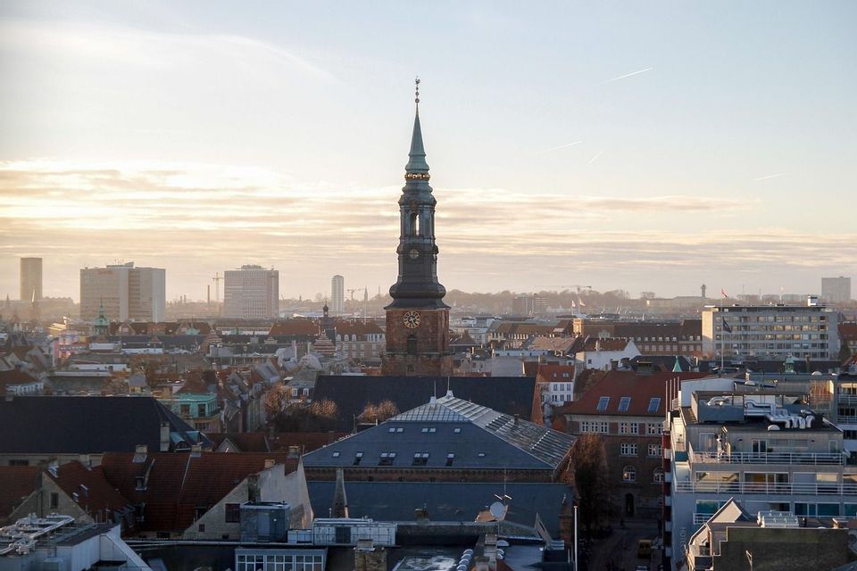 Eine Luftaufnahme von Dächern einer Stadt mit einem markanten Uhrenturm, der sich vor einem diesigen, hellen Himmel erhebt.