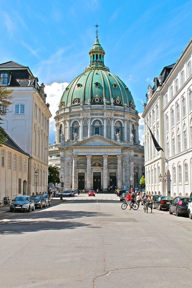 Eine große Steinkirche mit einer markanten grünen Kuppel steht an einem sonnigen Tag am Ende einer europäischen Stadtstraße.