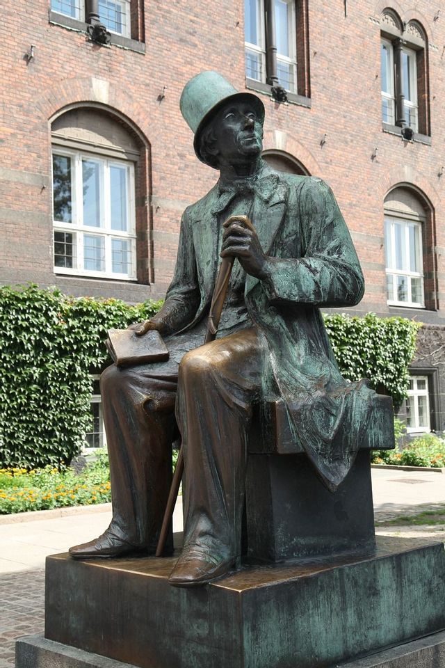 Eine Bronzestatue eines Mannes mit Zylinder, auf einem Sockel sitzend, ein Buch und einen Stock haltend, vor einem großen Backsteingebäude.