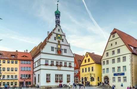 Un edificio blanco histórico con una torre de reloj de techo verde en una plaza de pueblo europeo, rodeado de casas de gablete coloridas.