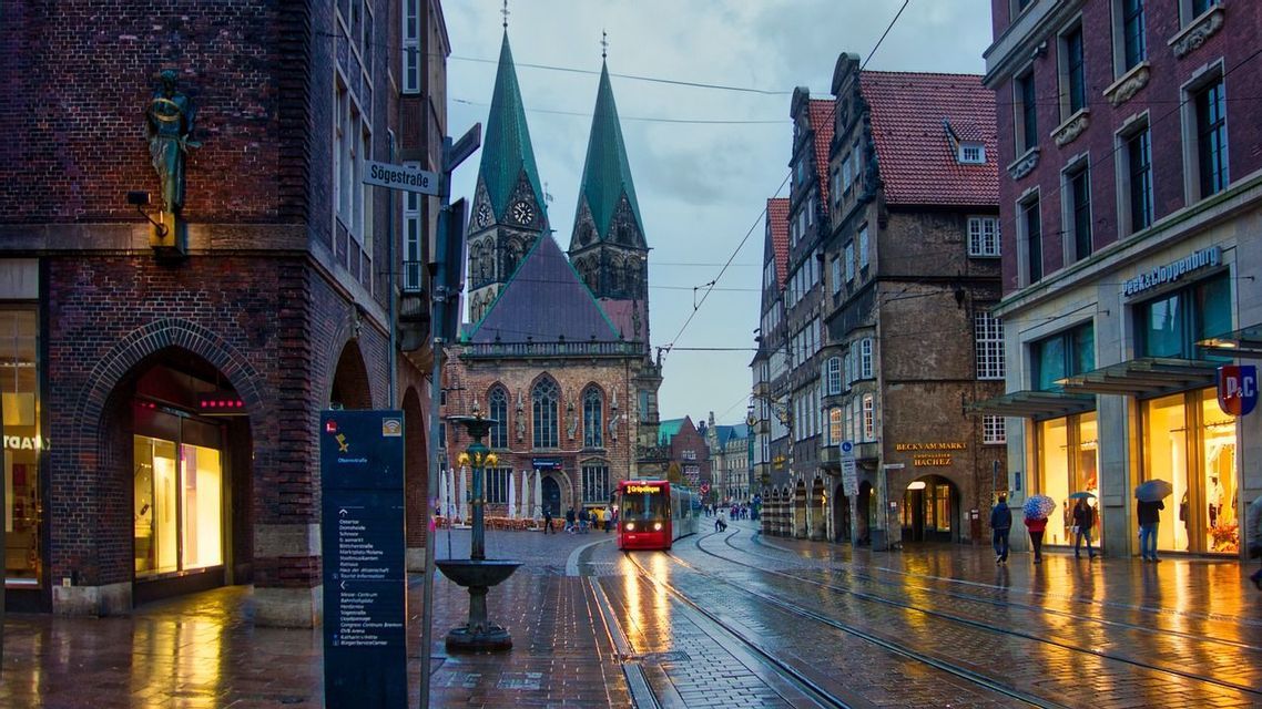 Un tranvía rojo viaja por vías mojadas a través de una calle de ciudad europea al anochecer, con una catedral de dos agujas al fondo.