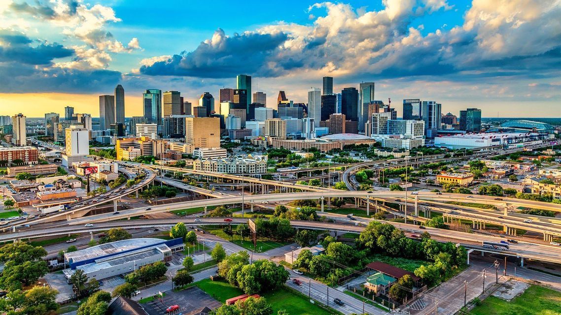 An aerial view of a modern city skyline with multiple highway interchanges in the foreground under a cloudy blue sky.